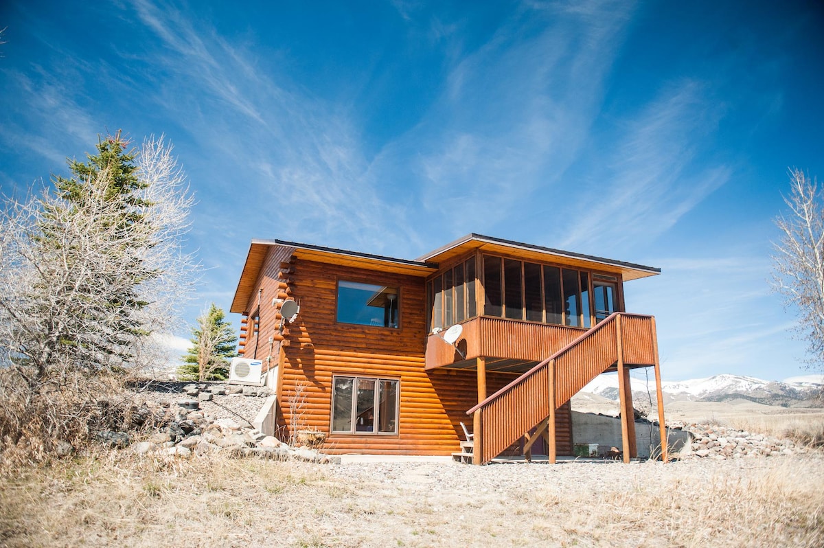 A modern cabin with wooden siding is surrounded by a natural landscape. The structure features multiple large windows, inviting light indoors. A staircase leads to a raised patio area, offering views of the surrounding scenery and mountains.