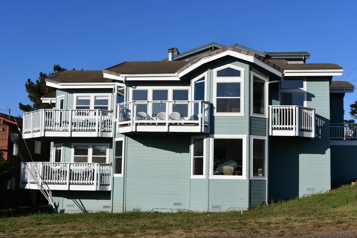 The two-story house displays a coastal architectural style with large windows and multiple balconies. The exterior is painted in soft blue, complemented by white railings. A spacious lawn is visible in the foreground, offering a serene outdoor space.