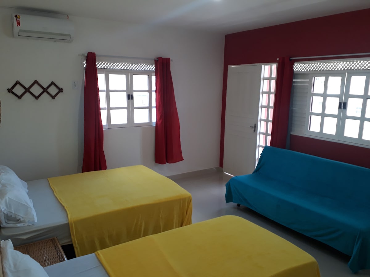 A well-lit living space displays two spacious beds covered with bright yellow blankets, alongside a blue sofa bed. Windows framed with white grilles allow natural light to filter in, while red curtains add a pop of color to the room's decor.