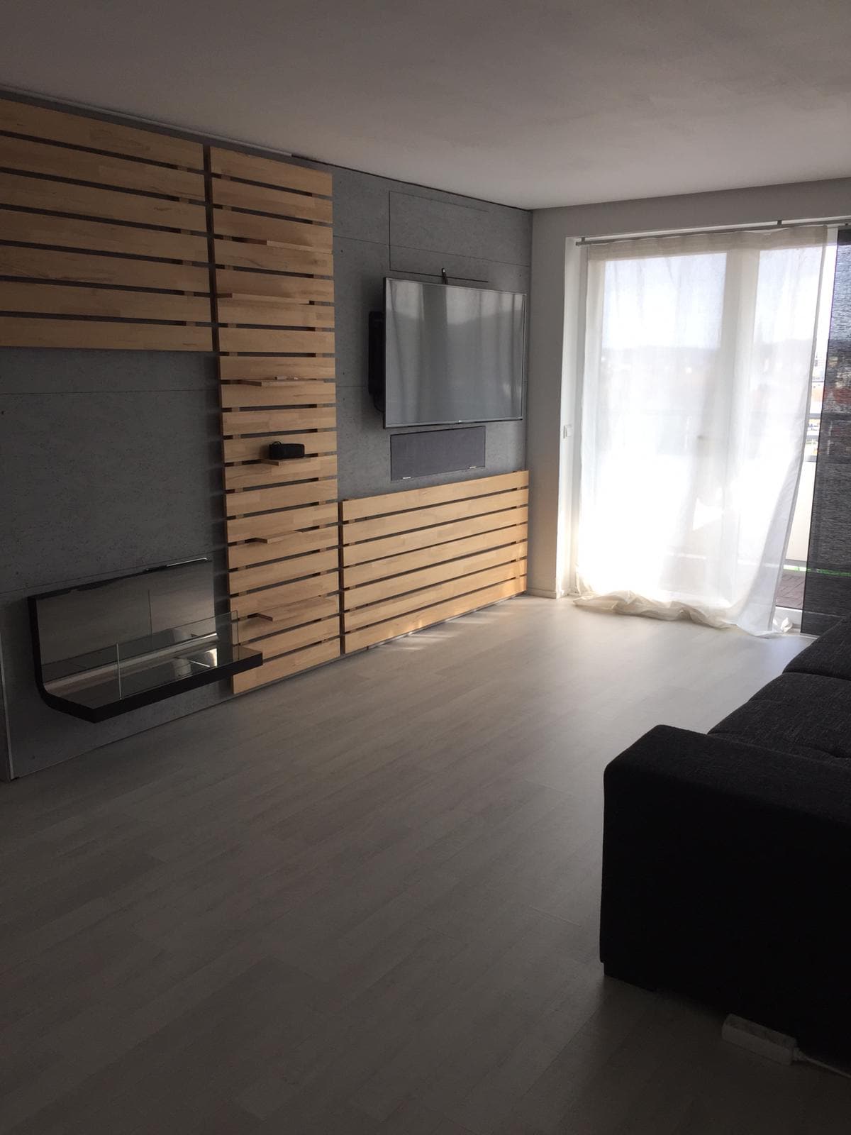 The living area features a modern design with wooden wall accents and a sleek wall-mounted television. A comfortable couch faces a minimalist fireplace, while natural light filters through sheer curtains, illuminating the light-colored flooring and creating a spacious atmosphere.