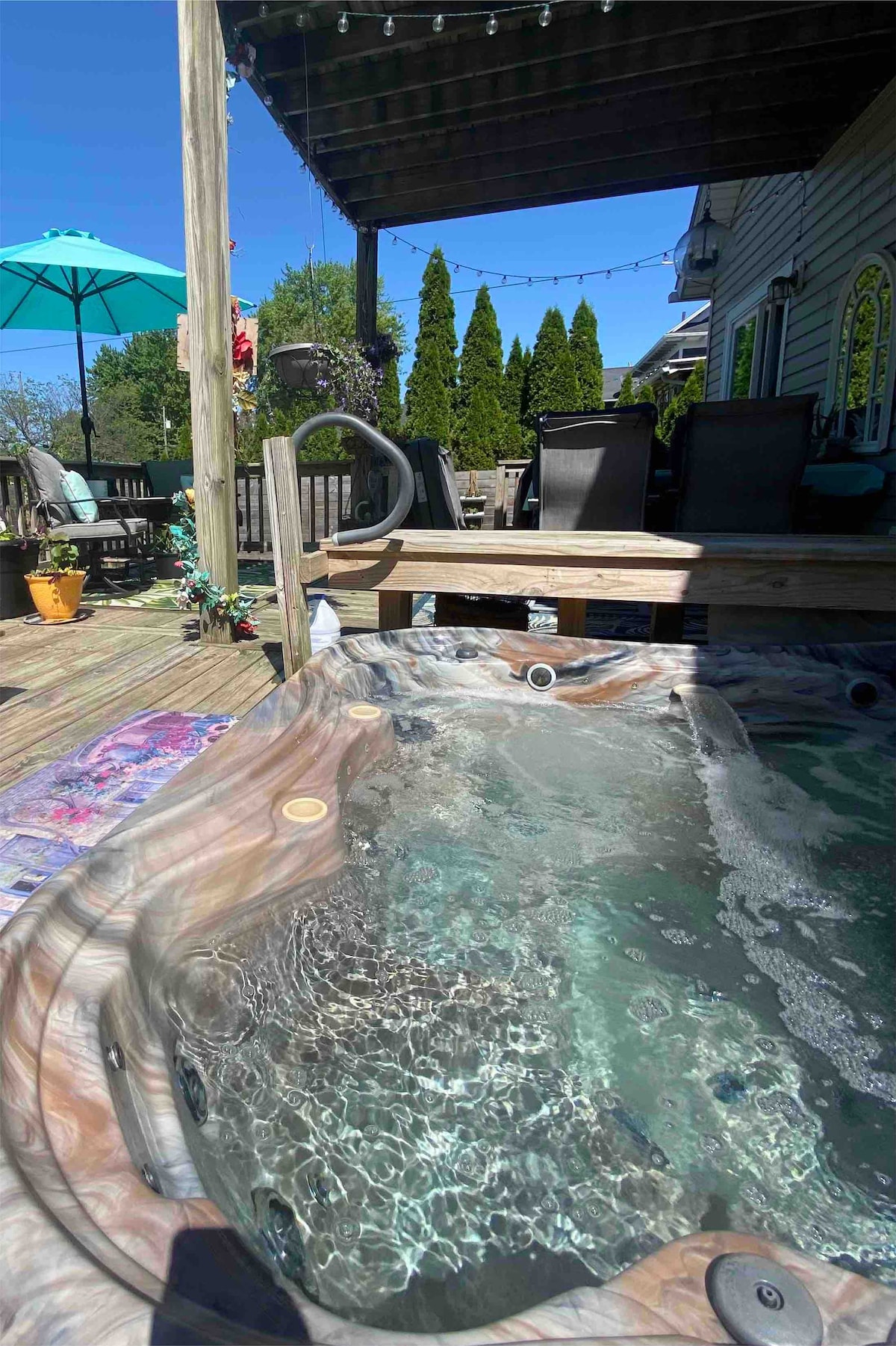 A built-in hot tub sits on the wooden deck, surrounded by a spacious outdoor area. The bubbling water is visible, creating a relaxing ambiance. Comfortable seating is arranged nearby under a bright umbrella, with greenery adding to the serene atmosphere.