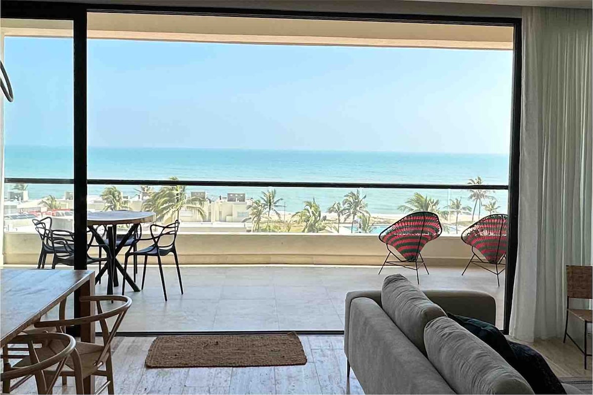 A bright living area showcases an expansive glass door that opens to a balcony with ocean views. A dining table for four and two chairs are positioned on the balcony, while a spacious sofa sits inside, inviting relaxation and enjoyment of the seaside scenery.