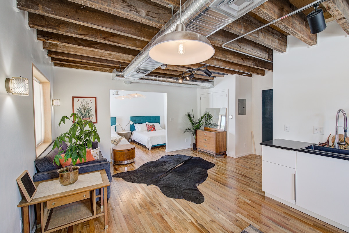 An open living area displays hardwood floors and exposed wooden beams. A cozy seating arrangement includes a sofa and coffee table, while a plush area rug adds texture. A spacious bed is visible in the background, complemented by minimalistic decor and natural light from a nearby window.
