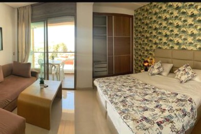 A cozy room features a spacious bed with patterned bedding, complemented by decorative pillows. A sofa and coffee table are arranged beside a large window, which offers access to a balcony. A floral wallpaper adds a touch of color to the serene atmosphere.