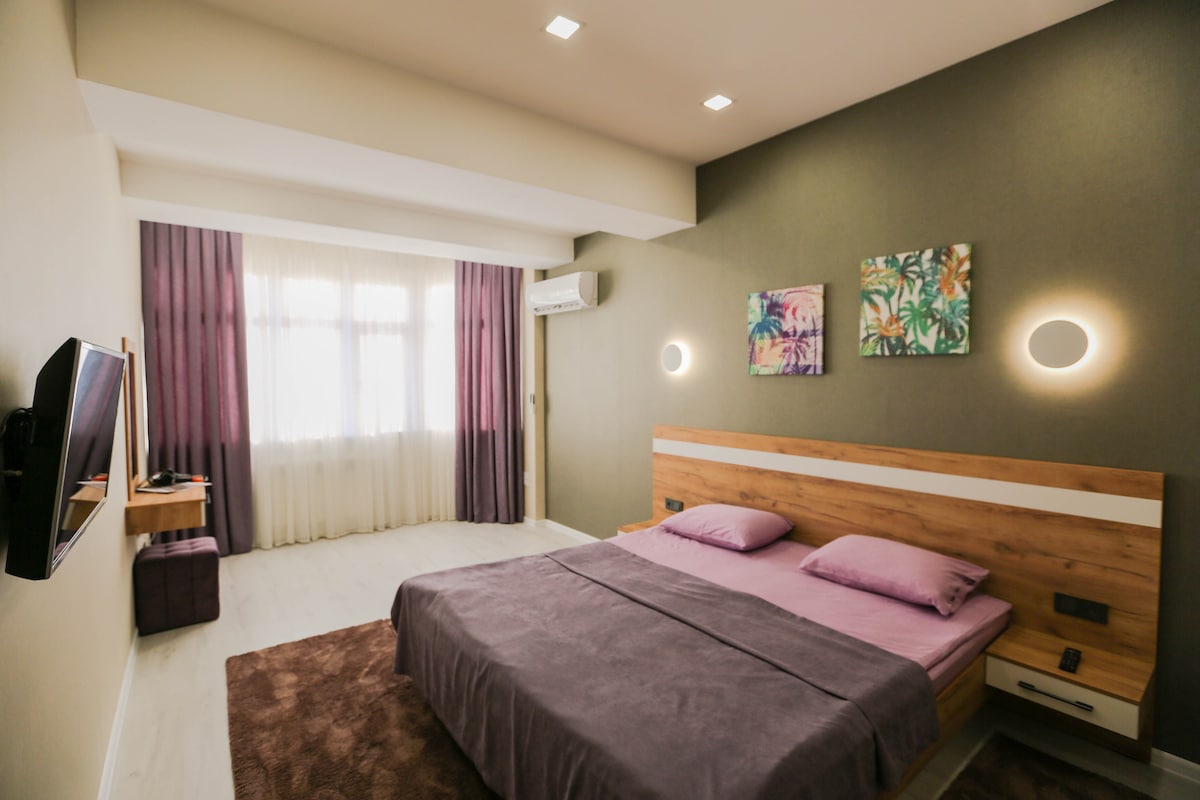A spacious room features a comfortable bed with pastel-colored linens, complemented by two colorful artworks on the wall. Natural light filters through the large window, with sheer curtains allowing a soft glow. A modern television is mounted across from the bed.