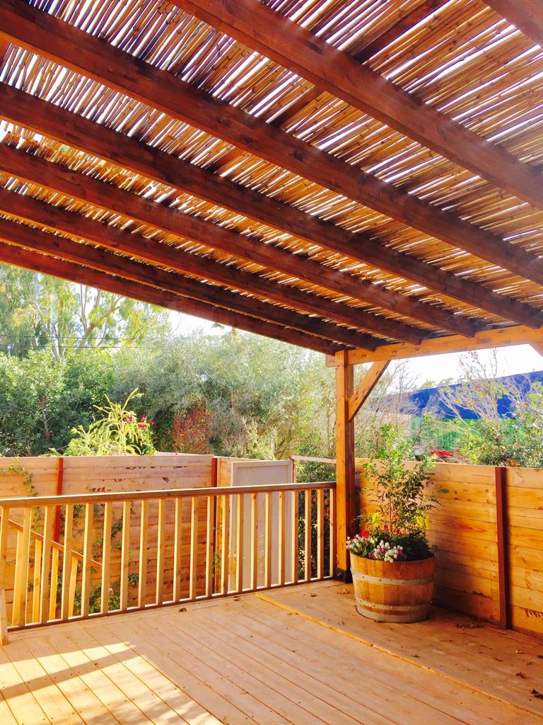 A large outdoor deck is presented, featuring a wooden structure with a slatted roof. Greenery surrounds the area, with potted plants adding natural elements. The deck is enclosed by wooden railings, creating a private space for relaxation.