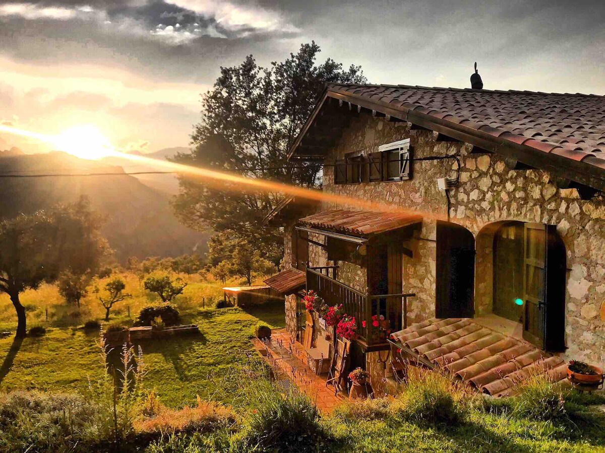 The historic Masia is set amidst lush greenery, showcasing a stone façade and wooden shutters. A sunbeam breaks through clouds, casting warm light on the exterior. Outdoor furniture and flower pots adorn the patio, with a view of the rolling hills and mountains in the background.