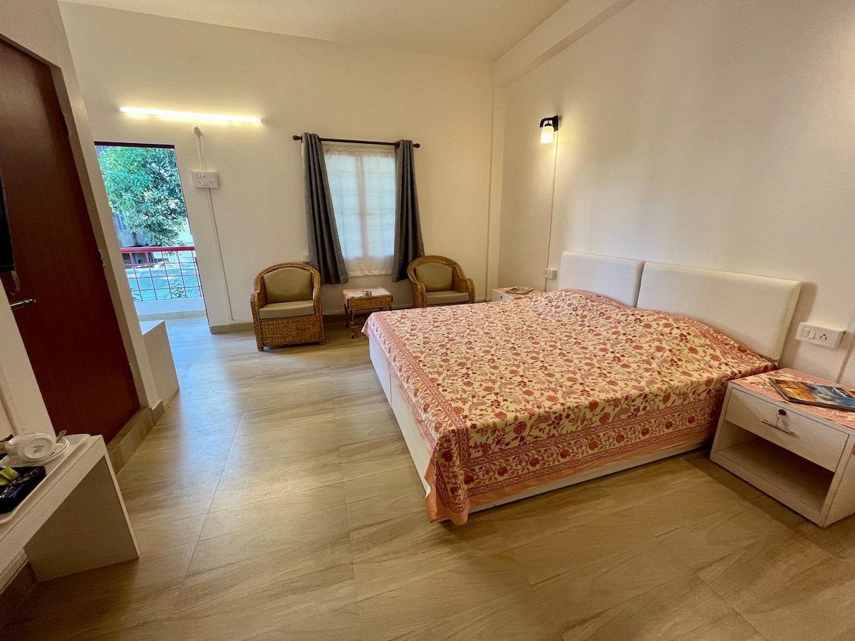 A spacious room features a large bed dressed in a floral bedspread. Two wicker chairs are positioned beside a window, allowing natural light to enter. Soft lighting enhances the room's brightness, while warm wooden flooring adds a soothing touch.