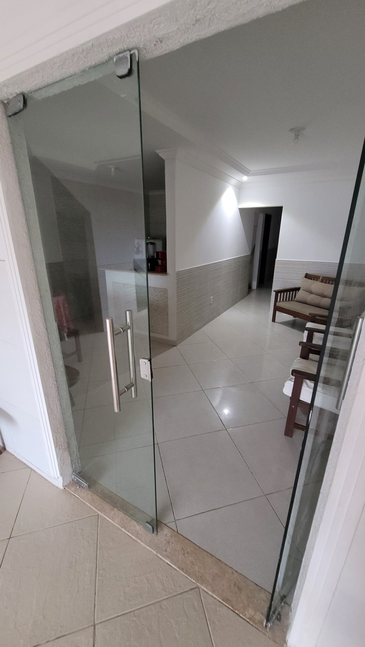 A clear glass door offers a view into a well-lit hallway featuring tiled flooring. Visible are two wooden chairs positioned near the entrance, with light reflecting off the glossy tiles, leading to a spacious area beyond.