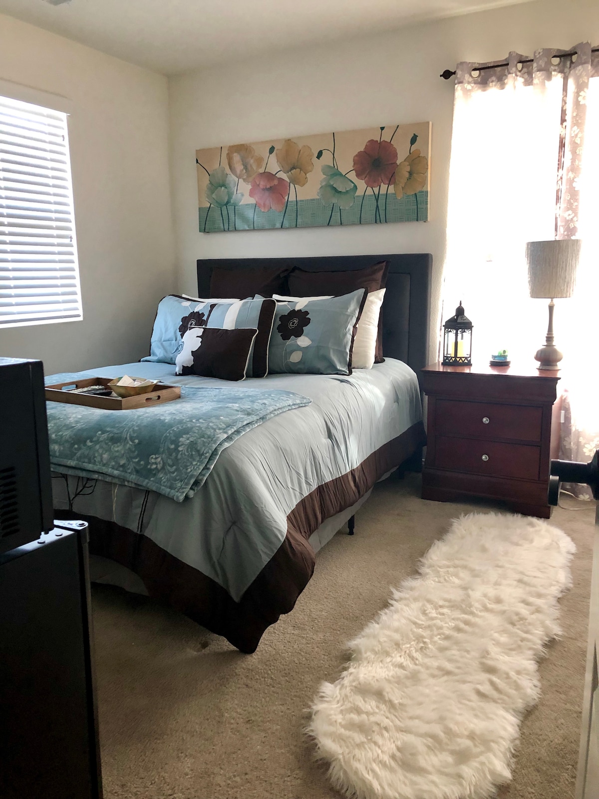 A bright and inviting bedroom features a spacious bed with light blue and brown bedding, complemented by decorative pillows. A small nightstand with a lamp sits beside the bed, and a wooden tray with items is placed on the bed. Soft carpeting covers the floor.