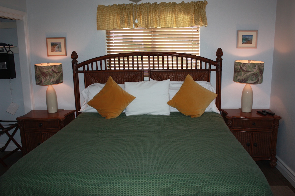 A spacious bed with a green coverlet is flanked by two bedside tables, each holding a lamp with patterned shades. Yellow accent pillows are placed against white bed linens. Soft light filters through a window, adorned with blinds and a decorative valance.