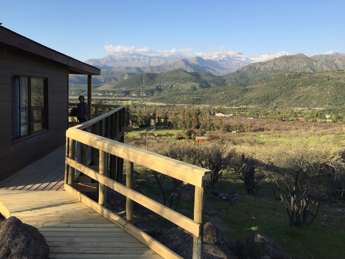 A wooden deck extends from the cabin, offering an unobstructed view of the surrounding mountains and lush landscape. The scene captures a serene atmosphere with a mix of greenery and distant peaks, framed by a clear blue sky.