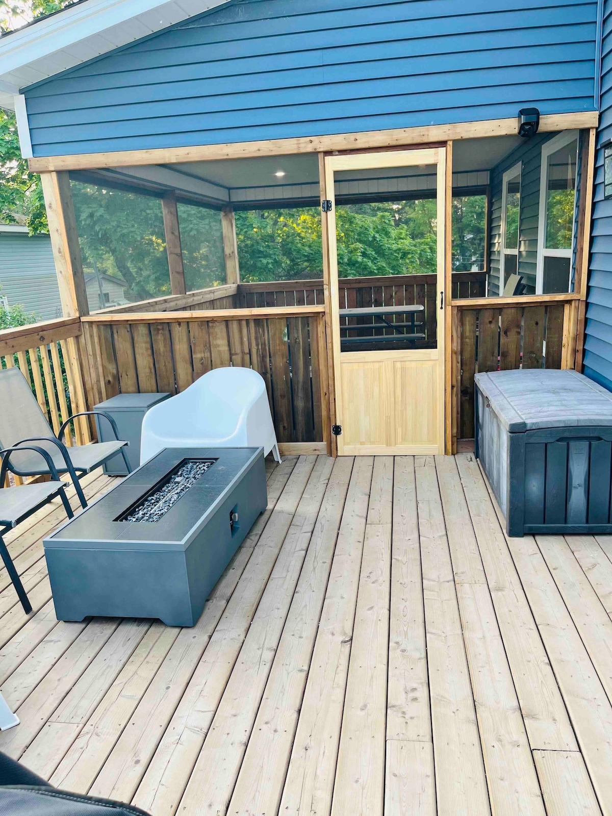 A screened-in porch is showcased with wooden walls and a door leading outside. Modern seating options include a white chair and gray outdoor fire table. The deck's wooden flooring complements a storage bench positioned against the wall, surrounded by a relaxing outdoor atmosphere.