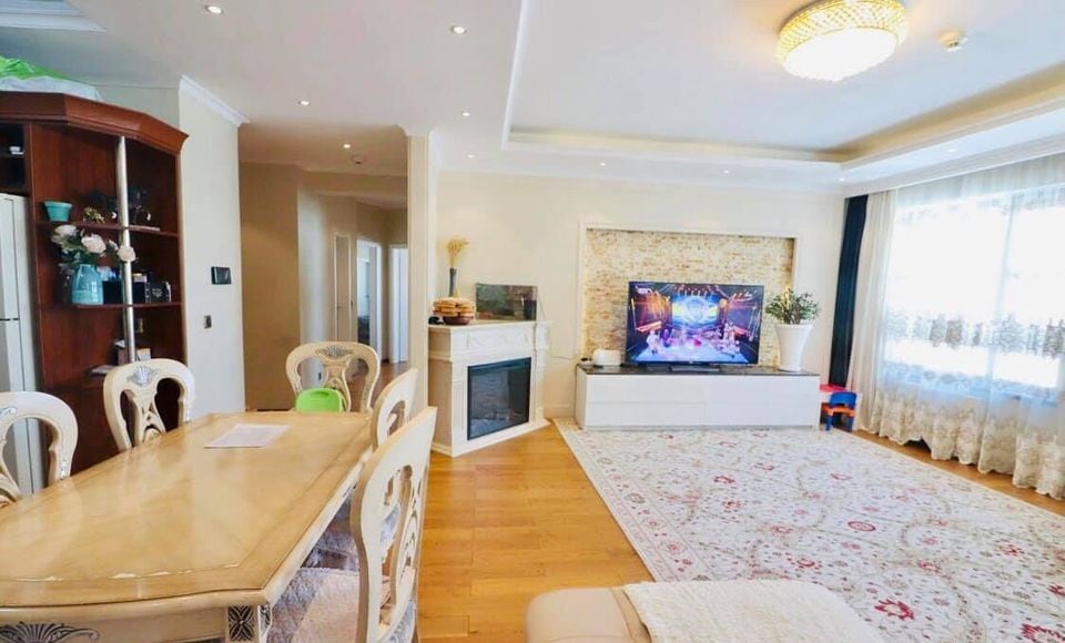 A bright living area features a comfortable seating arrangement centered around a television on a modern stand. A dining table is set against a wall cabinet, and a decorative fireplace adds style. Natural light filters through the sheer window coverings, illuminating the space.