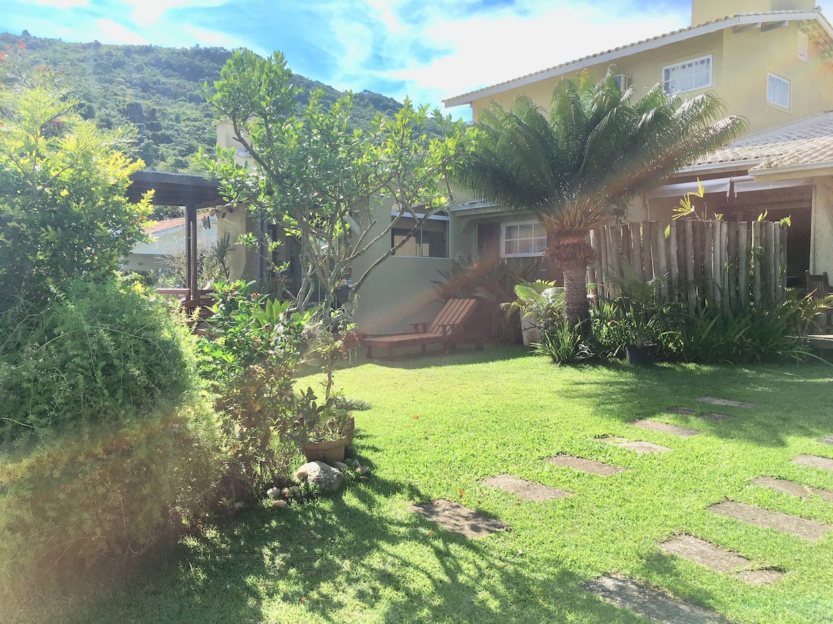A well-maintained garden area is visible, featuring lush green grass and various plants. Cobblestone pathways lead through the space, alongside a wooden deck and seating area beneath a pergola. A scenic backdrop of hills enhances the natural ambiance of the property.
