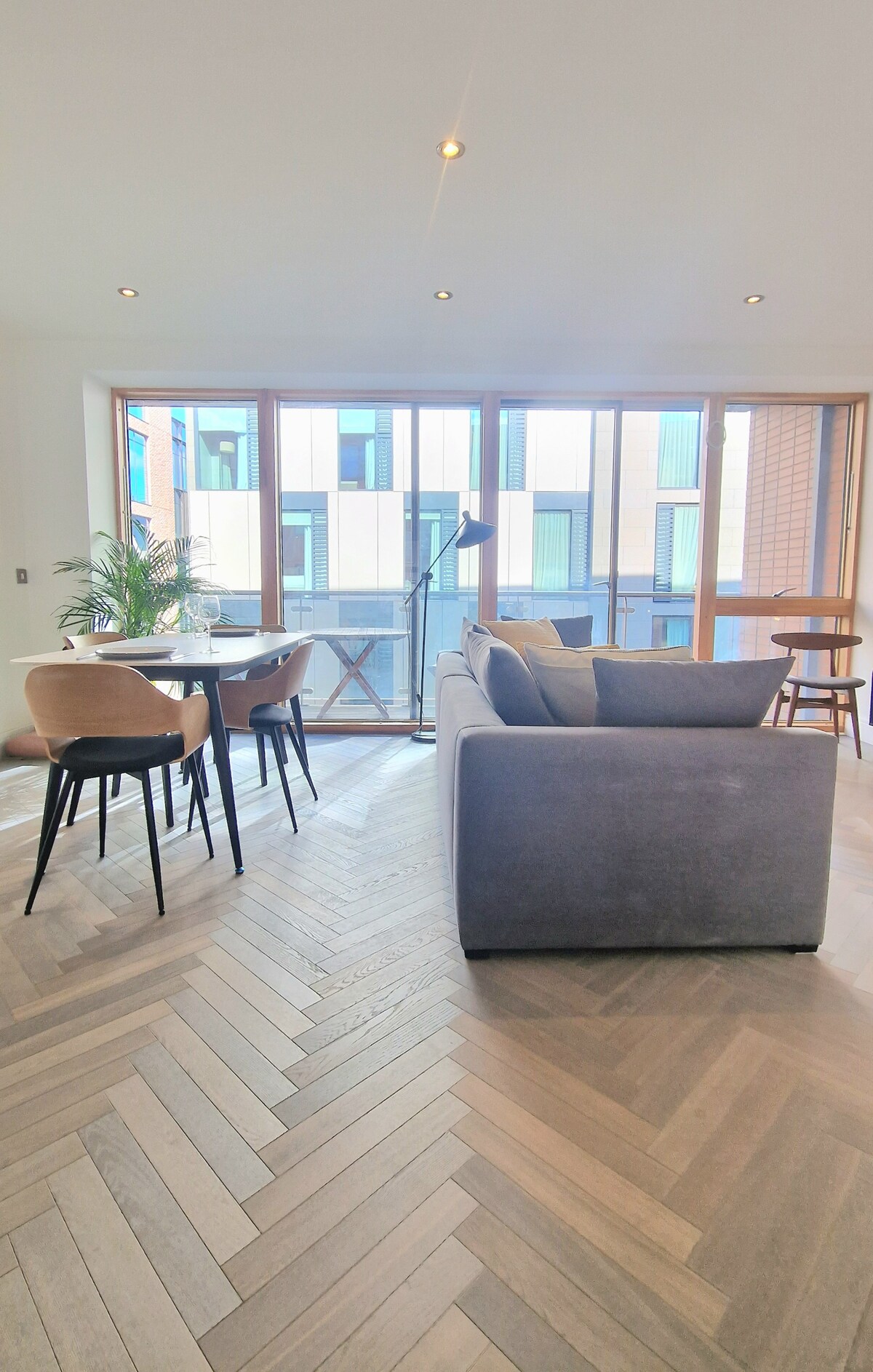 A modern living area is showcased with a spacious gray sofa facing large windows that provide natural light. A small dining table with four chairs sits nearby, alongside a sleek floor lamp. The herringbone-patterned wooden flooring enhances the contemporary aesthetic.