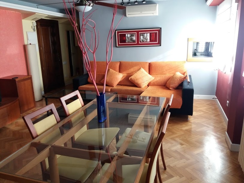 A spacious living area is equipped with a large orange sofa adorned with cushions. A glass dining table, surrounded by wooden chairs, offers seating for six. Parquet flooring is visible throughout, complemented by natural light entering from multiple windows.