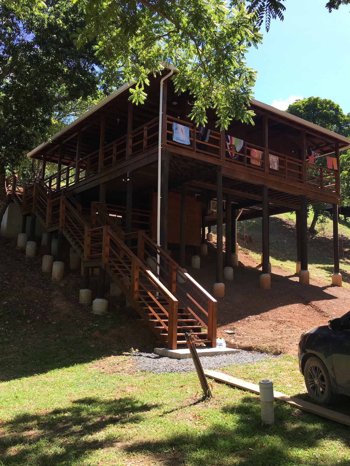 A wooden house elevated on stilts features a charming staircase leading to the entrance. Laundry items are hung on the balcony, while the surrounding greenery enhances the natural setting. A parked vehicle is visible in the foreground, with a pathway leading towards the house.