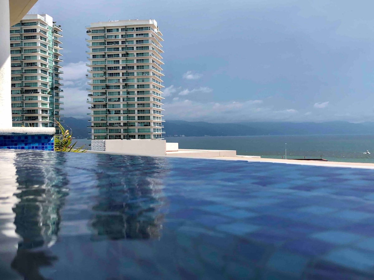 A serene view showcases the rooftop infinity pool, reflecting the sky and surrounded by lush greenery. In the distance, modern high-rise buildings stand alongside the coastline, with the ocean extending beyond, under a partly cloudy sky.