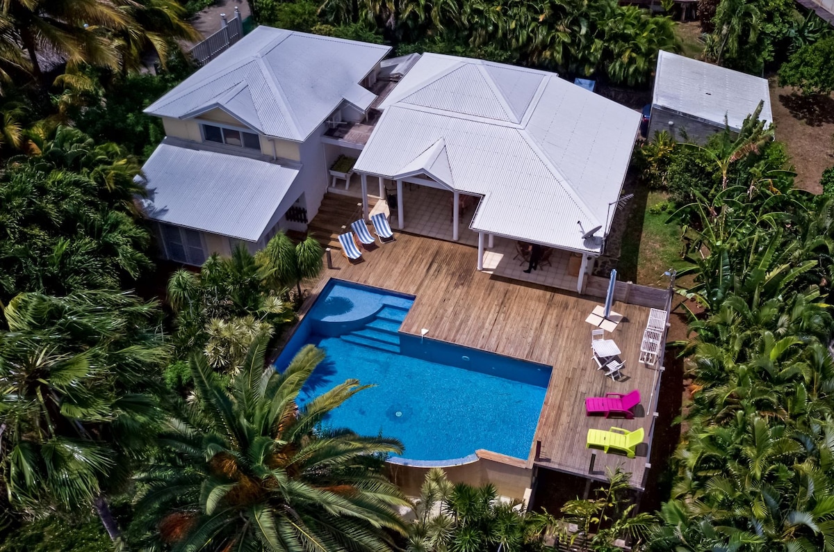 An aerial view of the villa showcases a spacious layout surrounded by lush greenery. The salt-water pool is framed by wooden decking, with sun loungers positioned strategically. The villa's roof and outdoor seating area are visible, indicating a blend of relaxation and comfort in a tropical setting.