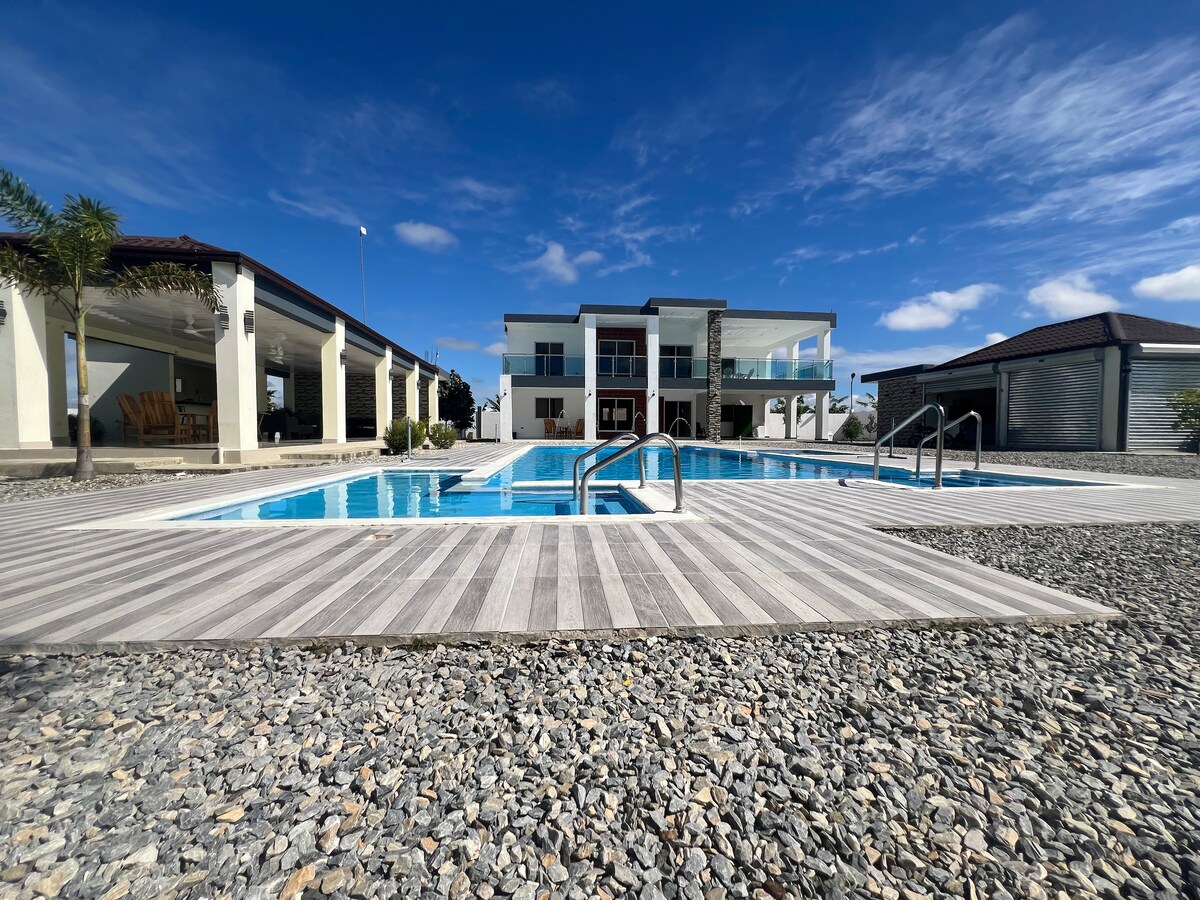 A well-maintained property features a clear swimming pool bordered by a wooden deck and smooth pebbles. The spacious two-story building with large windows overlooks the pool area, providing ample natural light and an inviting outdoor space. Lush palm trees are positioned around the property.