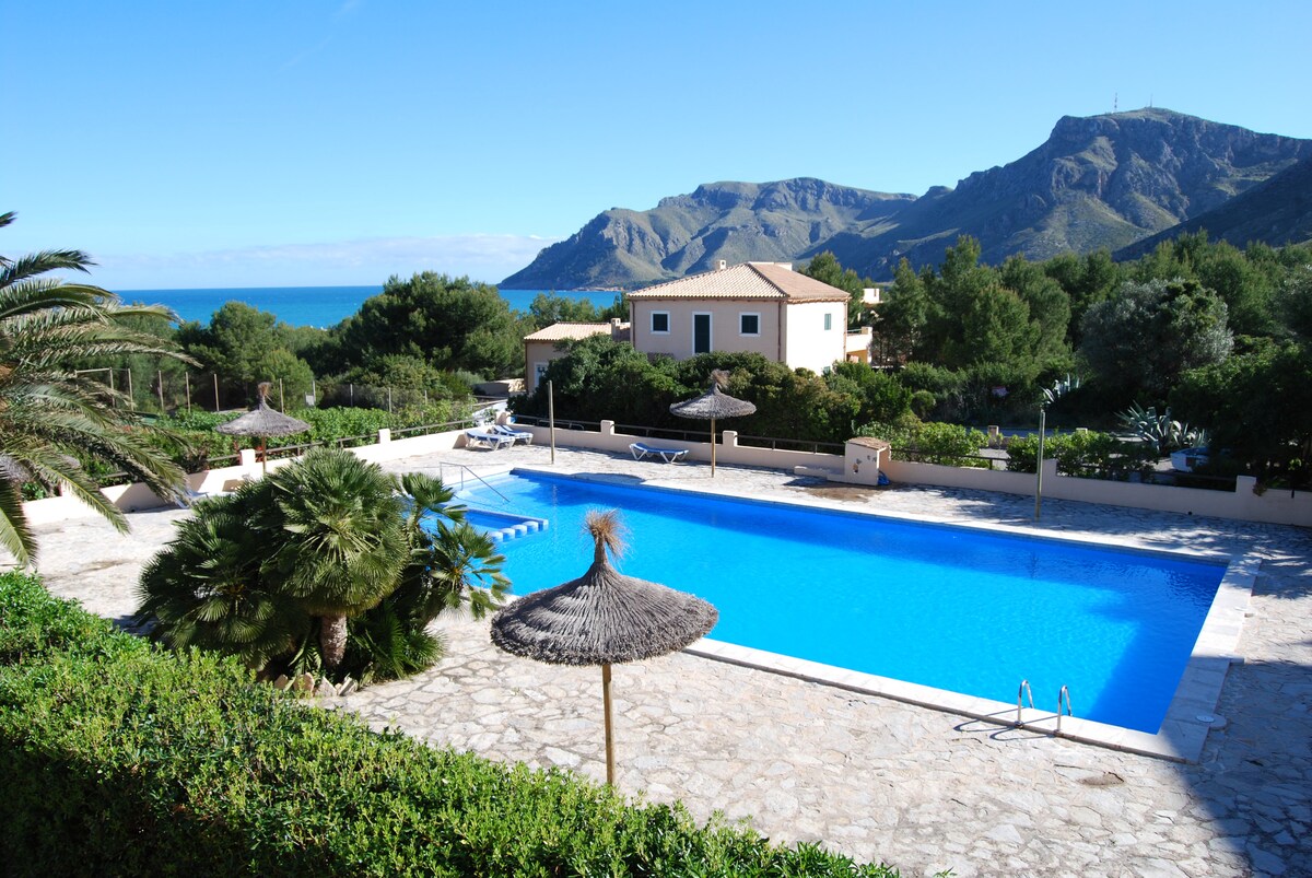 A large community swimming pool offers a refreshing blue expanse, surrounded by stone paving and lush greenery. Palm trees and sun umbrellas dot the area, providing shade. Scenic views of the mountains and sea create a serene backdrop for relaxation.