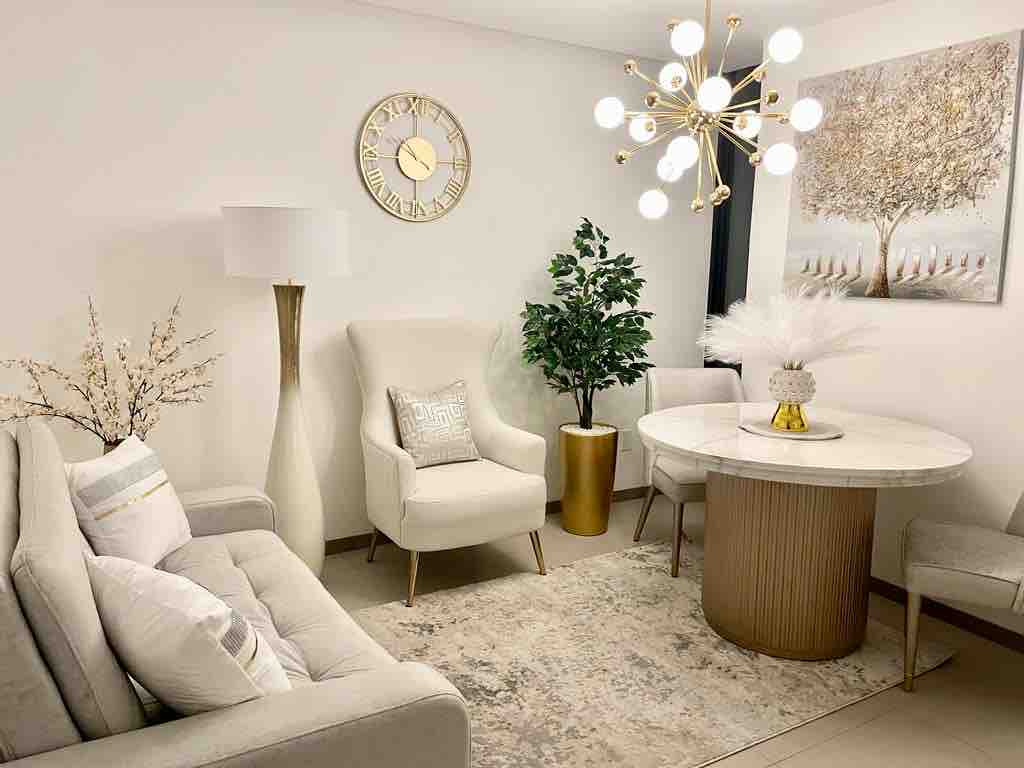 A cozy seating area is featured, including a light-toned sofa and an armchair. A round dining table is accented by a decorative centerpiece. A wall clock and a piece of art adorn the walls, while a floor lamp and indoor plant add warmth and life to the space.
