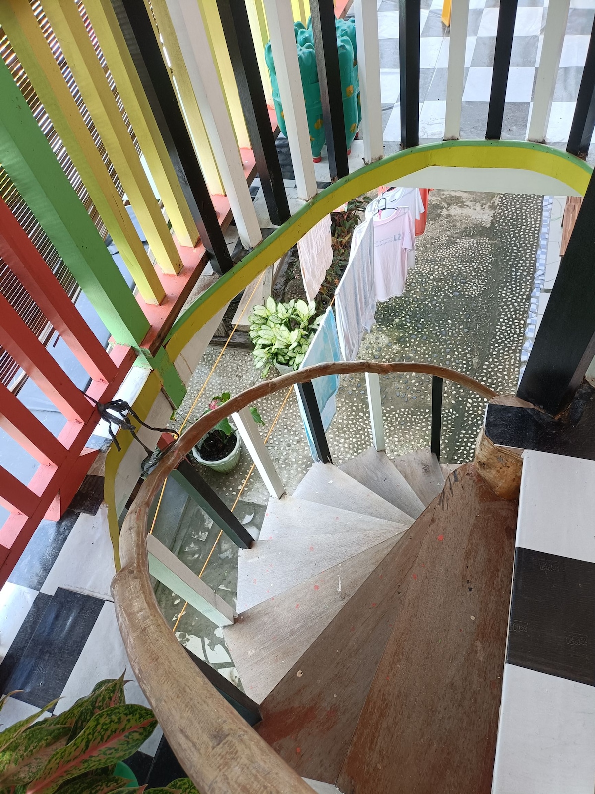The image captures a circular wooden staircase with a smooth handrail, winding down to the ground level. Colorful vertical slats frame the staircase, creating a vibrant contrast against the surrounding area, which features a patterned outdoor surface and greenery.