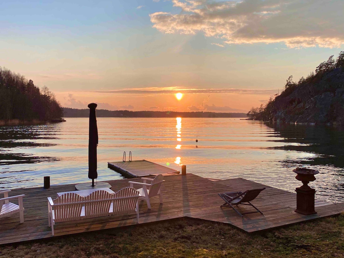 A serene waterfront scene features a wooden deck extending into calm waters. Two white lounge chairs and a black umbrella are positioned for relaxation. A gently setting sun casts a warm glow across the landscape, with subtle reflections visible on the water's surface.