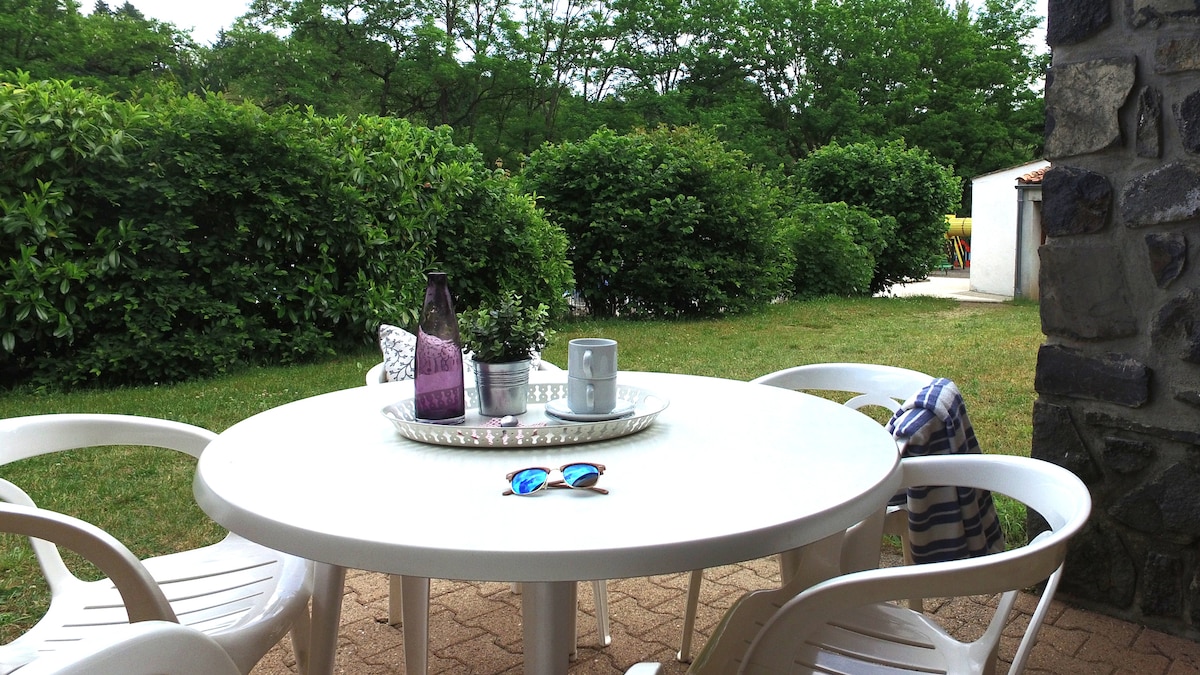An outdoor terrace is shown, equipped with a round white table and several stacked white chairs. A tray holds a cup and decorative items, while sunglasses rest on the table surface. Lush greenery and a well-kept lawn create a serene backdrop.