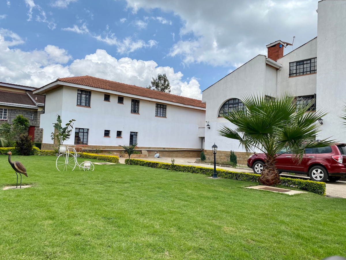 A well-maintained grassy area features manicured hedges and decorative white sculptures. Two buildings with distinct architecture stand side by side, with one showcasing large arched windows. A red vehicle is parked nearby, contributing to the inviting exterior ambiance.