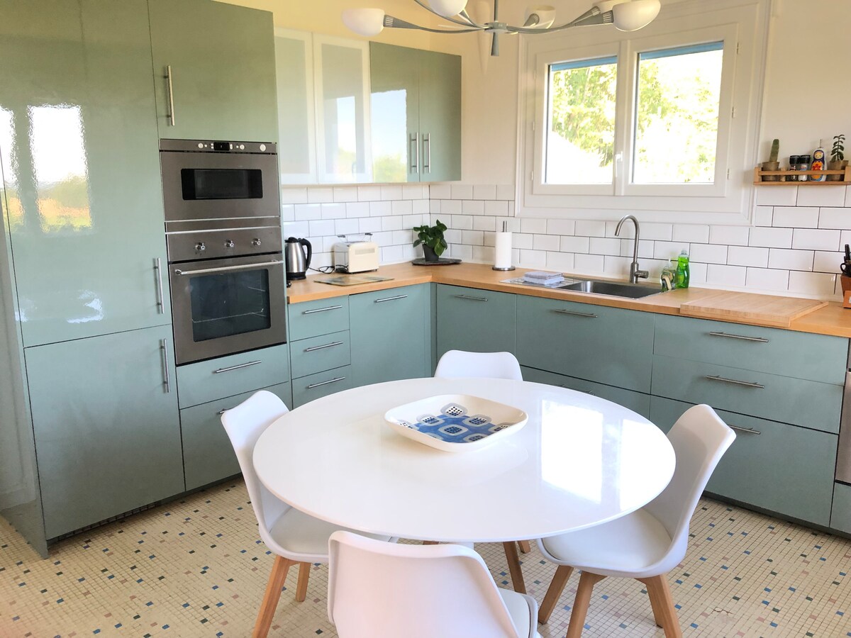 The kitchen features modern cabinetry in muted green tones and a light wood countertop. A round dining table with white chairs is centered in the space. Kitchen appliances, including an oven and kettle, are neatly arranged, complemented by a window that allows natural light in.