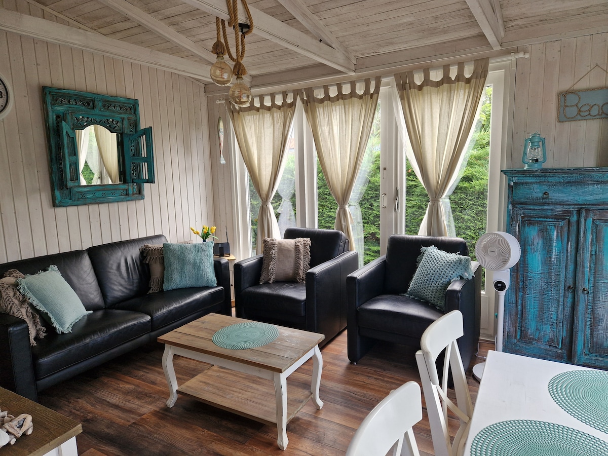 A welcoming living area features two black leather sofas and two armchairs, all adorned with decorative cushions. Natural light enters through large windows draped with light-colored curtains, illuminating a wooden coffee table and a dining table with white chairs. A decorative blue cabinet adds charm.