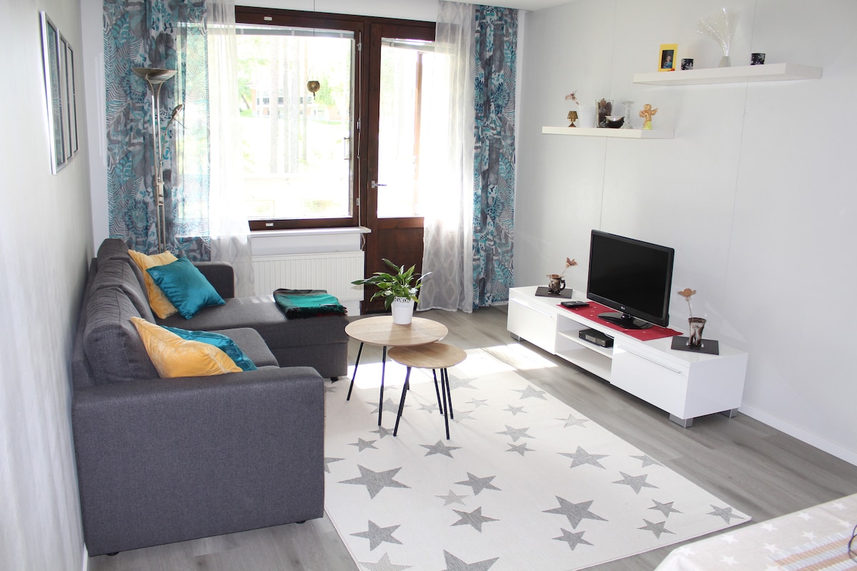 A living area features a gray sofa adorned with colorful pillows, paired with a round coffee table. Natural light filters through large windows, accented by sheer curtains. A television is positioned on a sleek white media console, with decorative items arranged on a nearby shelf.