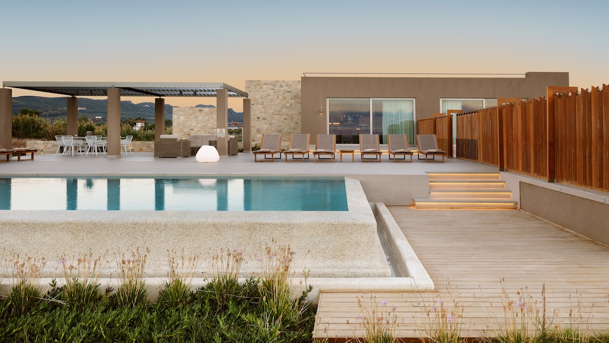 An infinity pool reflects the evening sky, bordered by sun loungers and a wooden deck. A shaded outdoor dining area is visible in the background, accompanied by a sturdy stone structure that adds modern appeal to the landscape.