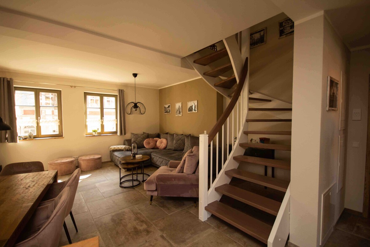 A spacious living area is observed with a large sectional sofa and cozy poufs arranged around a circular coffee table. A dining table is positioned nearby. A graceful spiral staircase leads to the upper level, and ample natural light fills the room through multiple windows.
