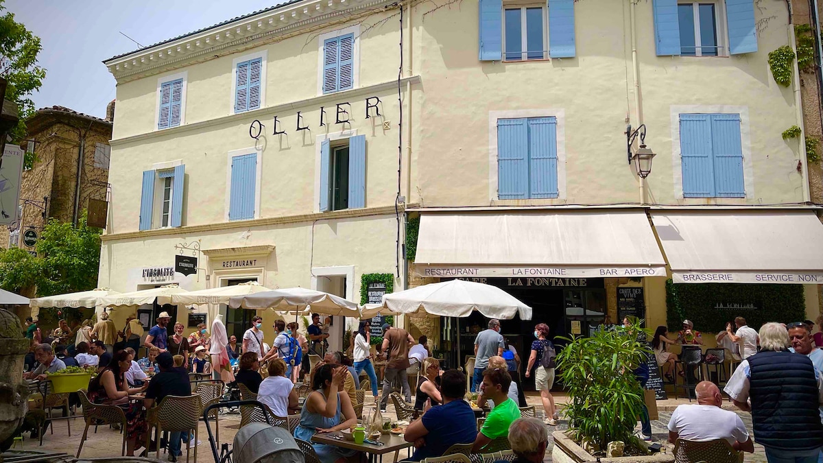 A vibrant square is filled with people enjoying outdoor seating at various café tables. Sunlight brightens the scene, highlighting the historic building facade with blue window shutters and a prominent sign that reads 'Ollier.' Umbrellas provide shade for patrons gathered under them.