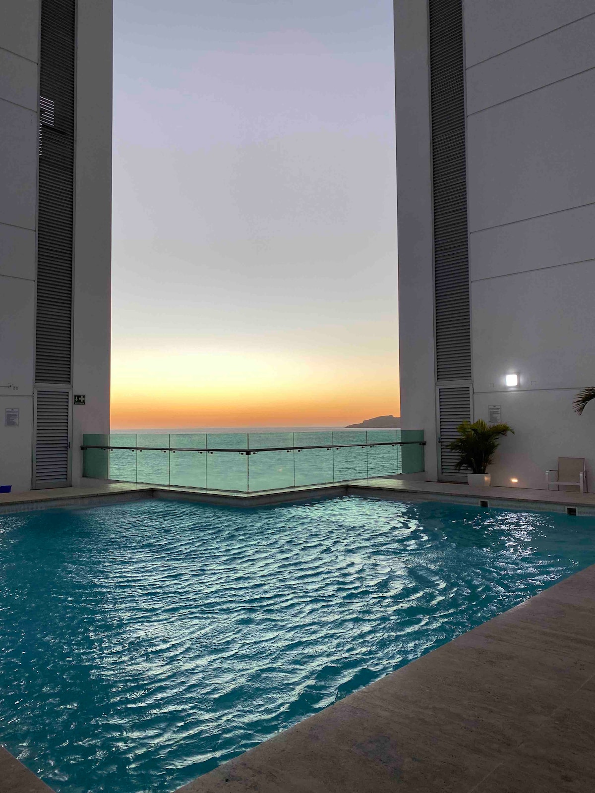 A swimming pool is situated between two modern building structures, reflecting the calm turquoise water. The horizon features a soft gradient of pastel colors as the sun sets, while the ocean glistens in the background, providing a serene view.