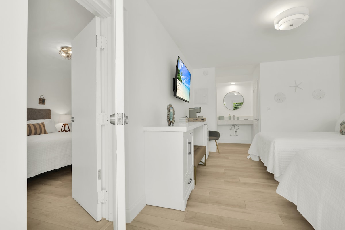 A light-filled hotel room is visible through an open doorway. Two beds with white bedding are placed against a neutral wall. A small desk area is situated beside a mounted TV, with an adjoining bathroom partially visible in the background.
