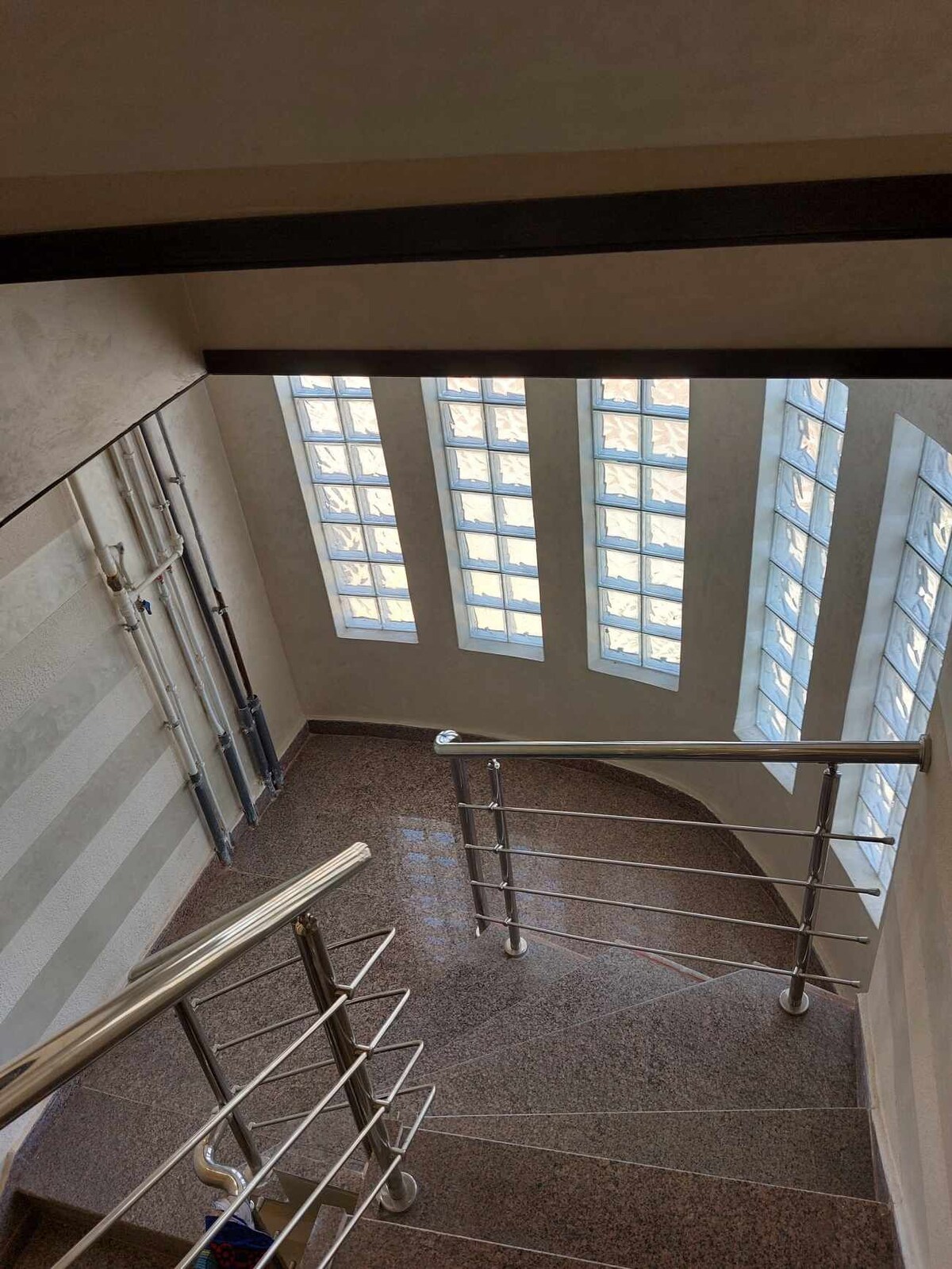 A brightly lit stairwell features a modern railing and a series of narrow, vertical windows that allow natural light to flood the space. The polished floor reflects the light, enhancing the spacious feel of the area.