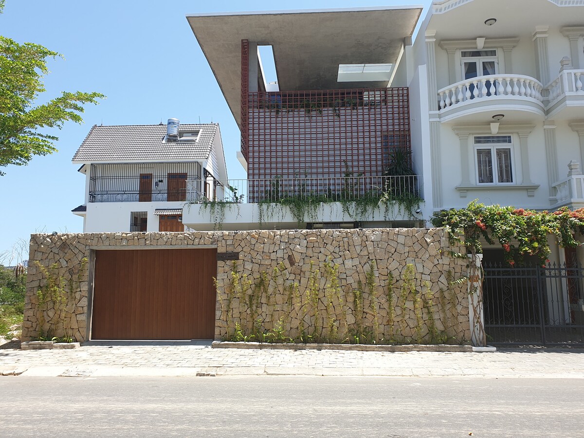 A modern building with a unique roof design is presented, featuring a stone wall with a wooden garage door at the front. A balcony with greenery is visible on the upper floor, complementing the structure's contemporary appearance.
