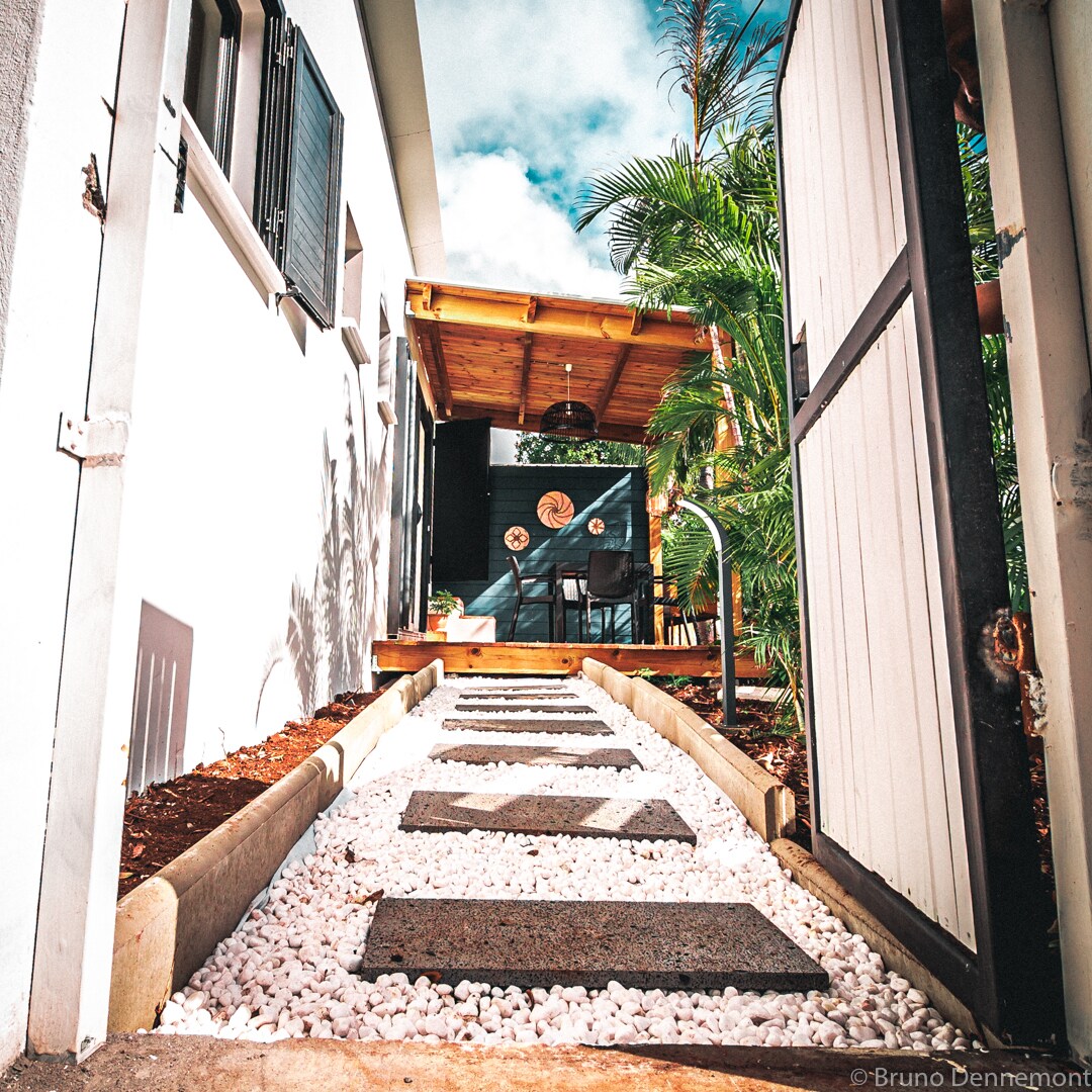 A pathway lined with rectangular stone slabs and white gravel is seen leading towards a wooden awning. Lush greenery frames the entrance, with a glimpse of seating visible beneath the awning in the background.