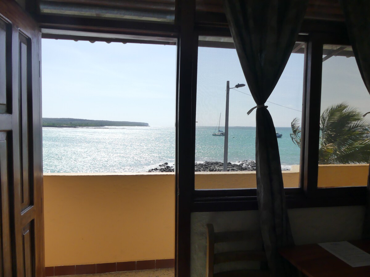 A scenic view of the ocean is framed by large windows, with shimmering water reflecting sunlight. The coastline features a distant shoreline and a sailboat. A wooden chair and table are visible in the foreground, complementing the bright atmosphere.
