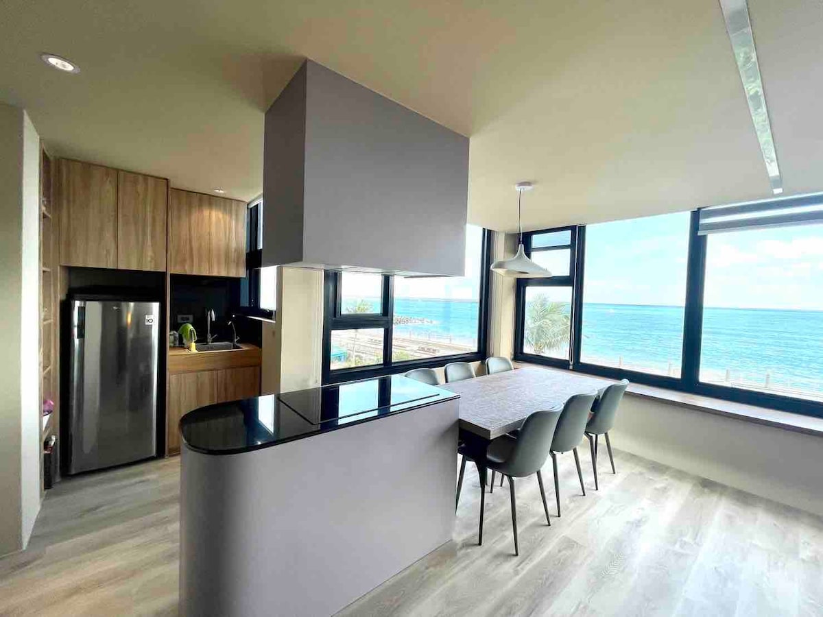 The kitchen features modern appliances and a spacious layout. A large dining table with several chairs overlooks the ocean through expansive windows. The contemporary kitchen island offers additional seating and counter space, creating a functional area for meal preparation and dining.