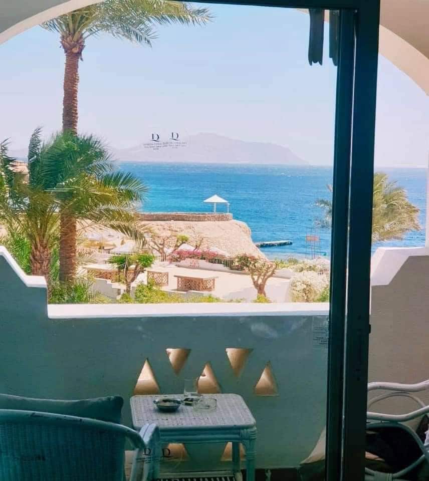 A balcony view features a tranquil sea foreground and distant mountains. Palm trees frame the scene, and a white canopy is visible near the water. A small table and chairs are positioned on the balcony, inviting relaxation in the coastal atmosphere.