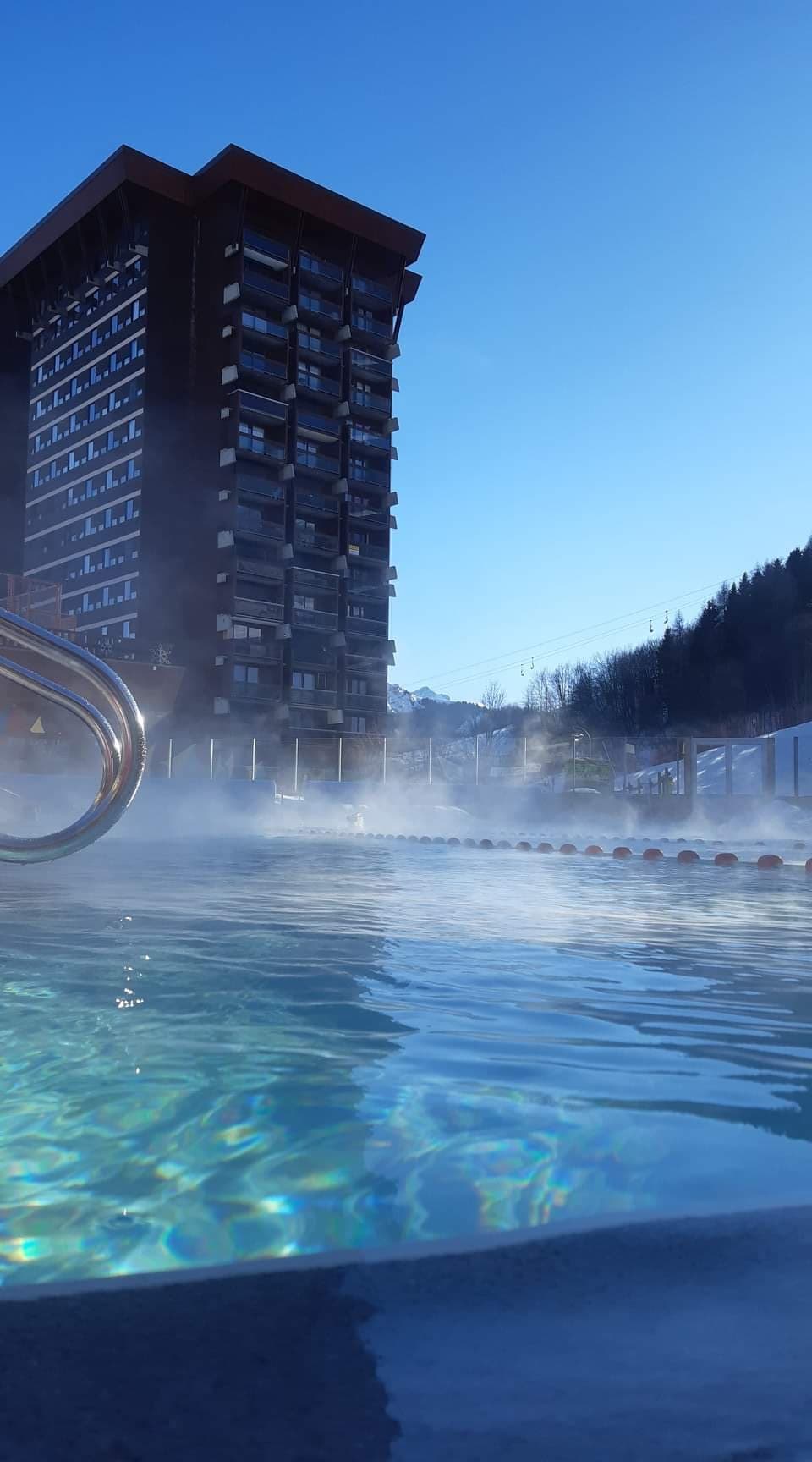 A serene outdoor swimming pool is depicted with gentle steam rising from the water. The architecture of a tall building is visible in the background, framed by a clear blue sky and distant mountains. The scene conveys a tranquil atmosphere.