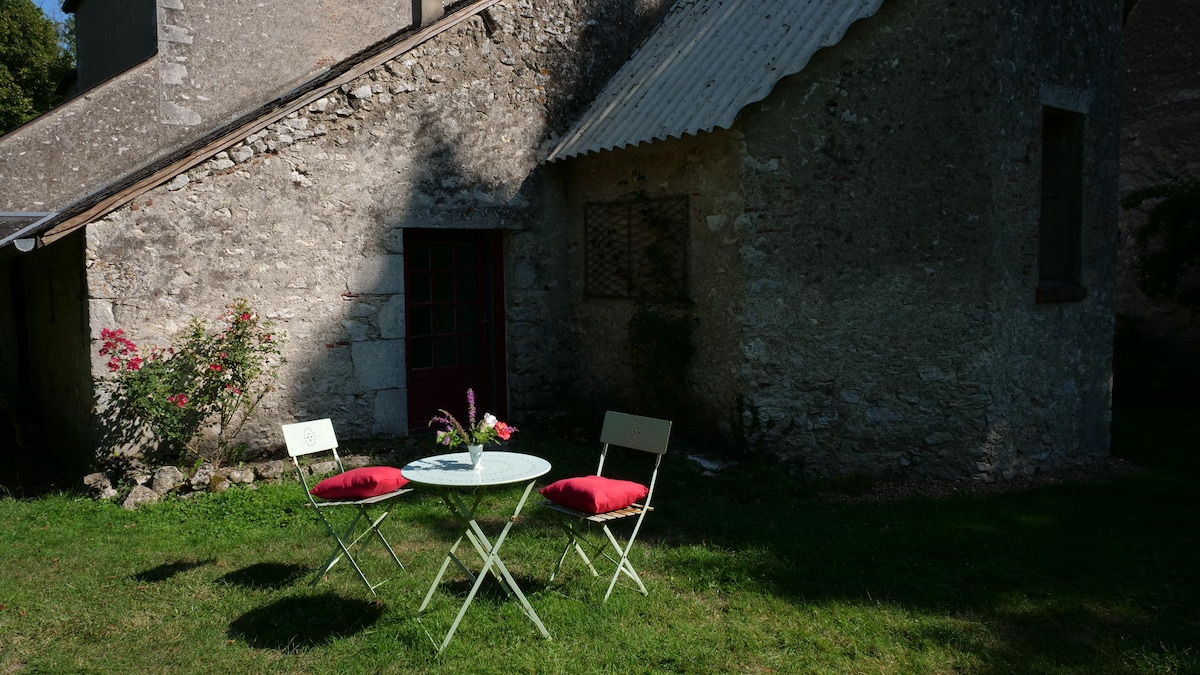 An outdoor seating area is presented in front of a rustic stone building. A small round table is set with a vase of flowers, accompanied by two folding chairs, featuring red cushions. Lush green grass surrounds the area, enhancing the peaceful setting.