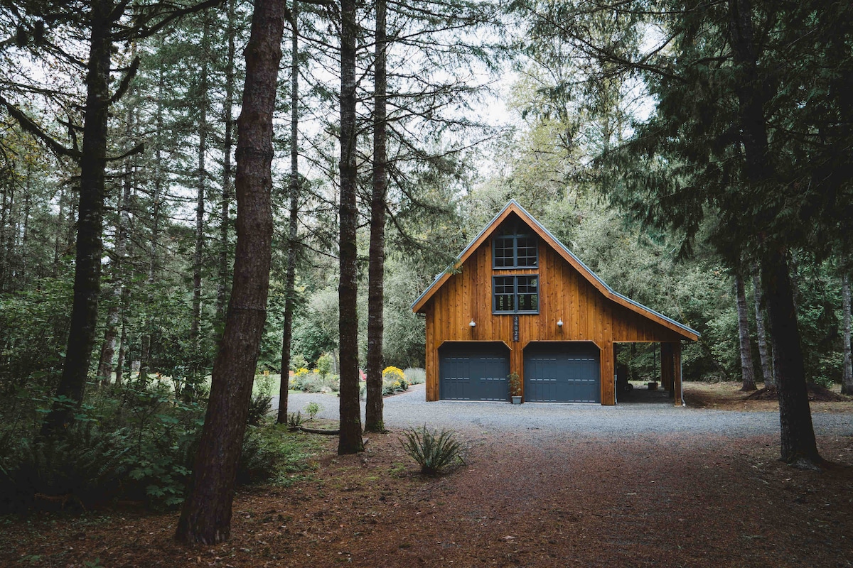 The timber frame structure is set amidst a forest, with a gravel driveway leading to the dual garage doors. Large windows allow natural light to fill the upper level, framed by tall trees creating a serene and secluded atmosphere.