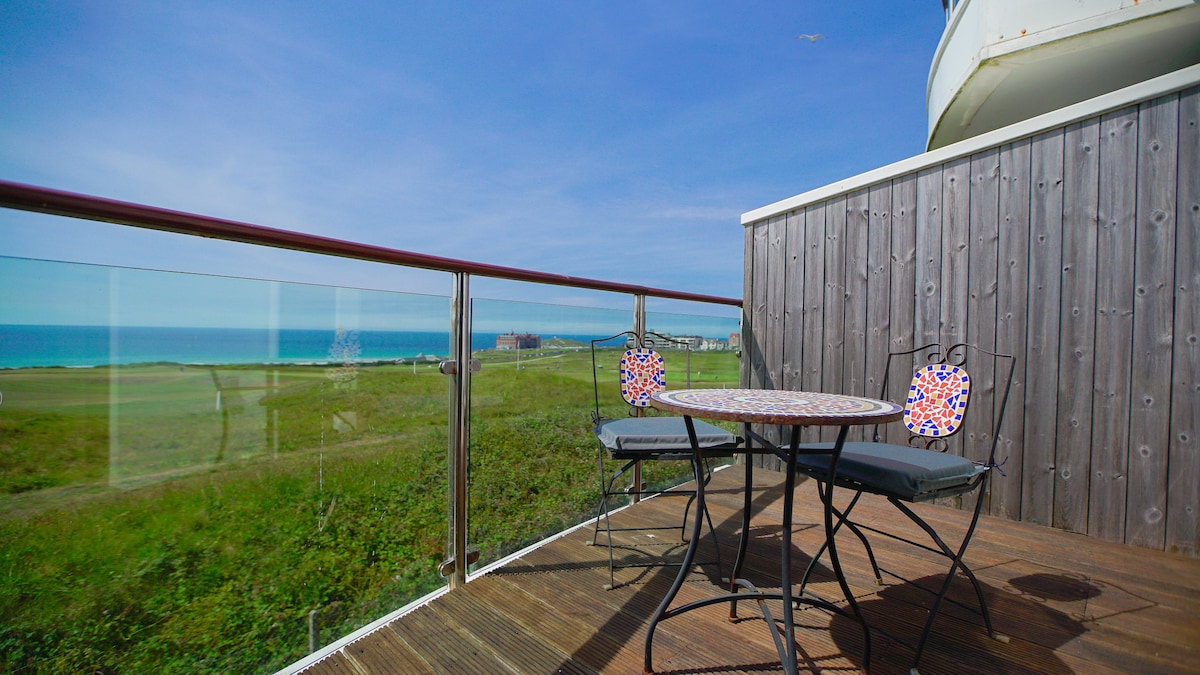 A small balcony overlooks scenic views of the sea and green landscape. A round table is set with two chairs, featuring decorative patterns. The glass railing allows for unobstructed views, while the wooden flooring complements the natural surroundings.
