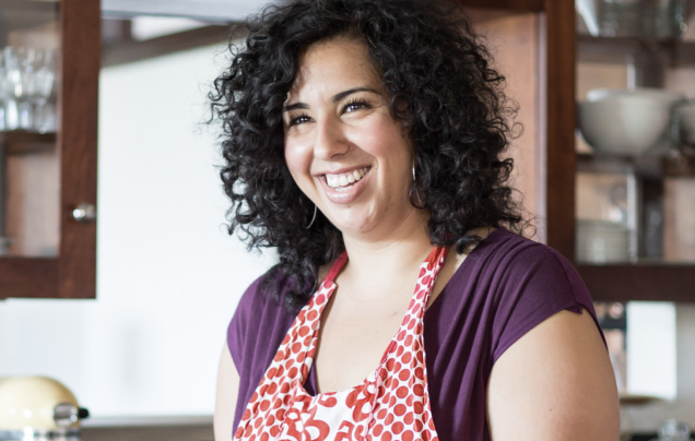 A woman with curly hair stands in a kitchen. She’s grinning and wearing a brightly colored apron.